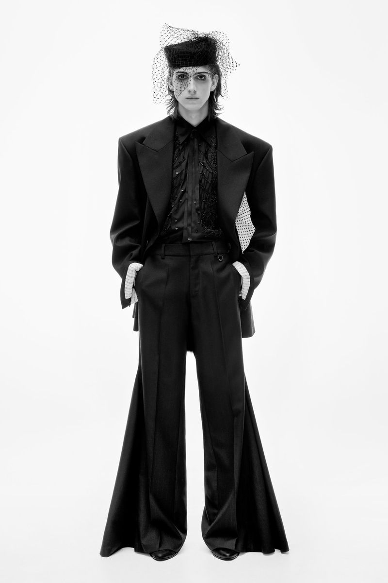 Model poses in a black, oversized suit with flared pants and a veiled hat against a bright white backdrop in a studio fashion shot.