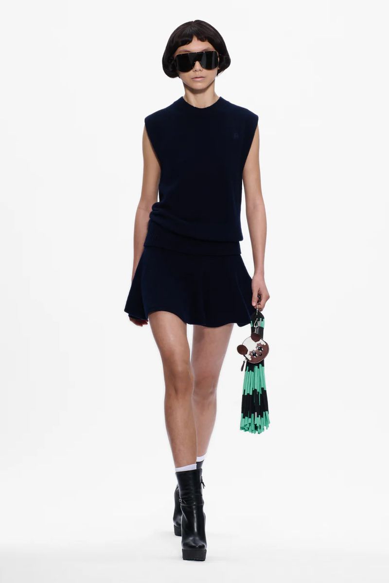 Fashion model on runway wearing a navy vest and matching short skirt with black platform boots and unique novelty handbag.
