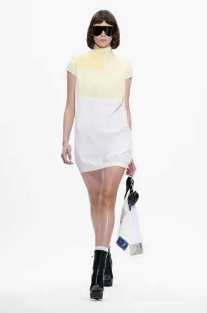 A slender model walks the runway in a two-tone mini dress, futuristic sunglasses, platform boots, and holding a draped textile.