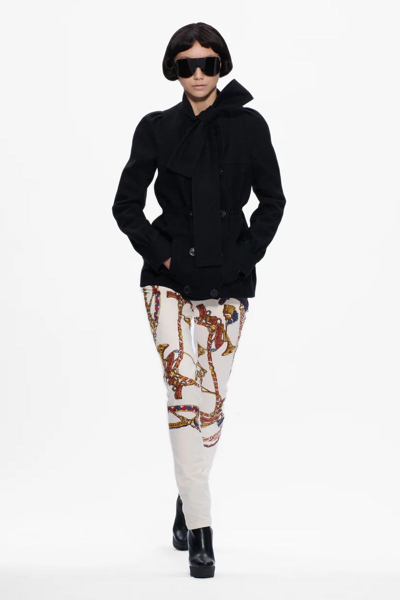 Runway fashion presentation featuring an adult female model wearing sunglasses, black jacket with scarf, and printed pants against white.