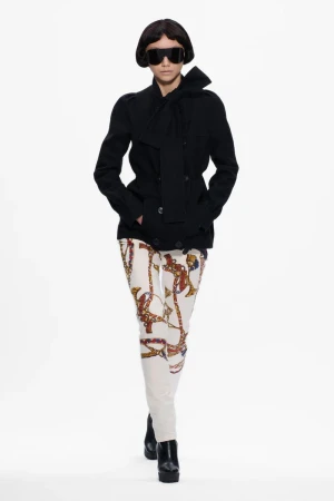 Runway fashion presentation featuring an adult female model wearing sunglasses, black jacket with scarf, and printed pants against white.