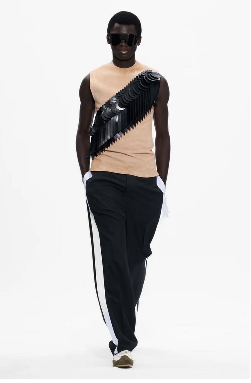 Runway fashion model walks on white in beige top with black sculptural accent, dark sunglasses, and black-and-white track pants.