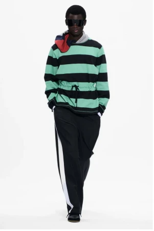 A runway shot of a man modeling a striped hooded jacket and black pants with a white stripe against a plain white background.