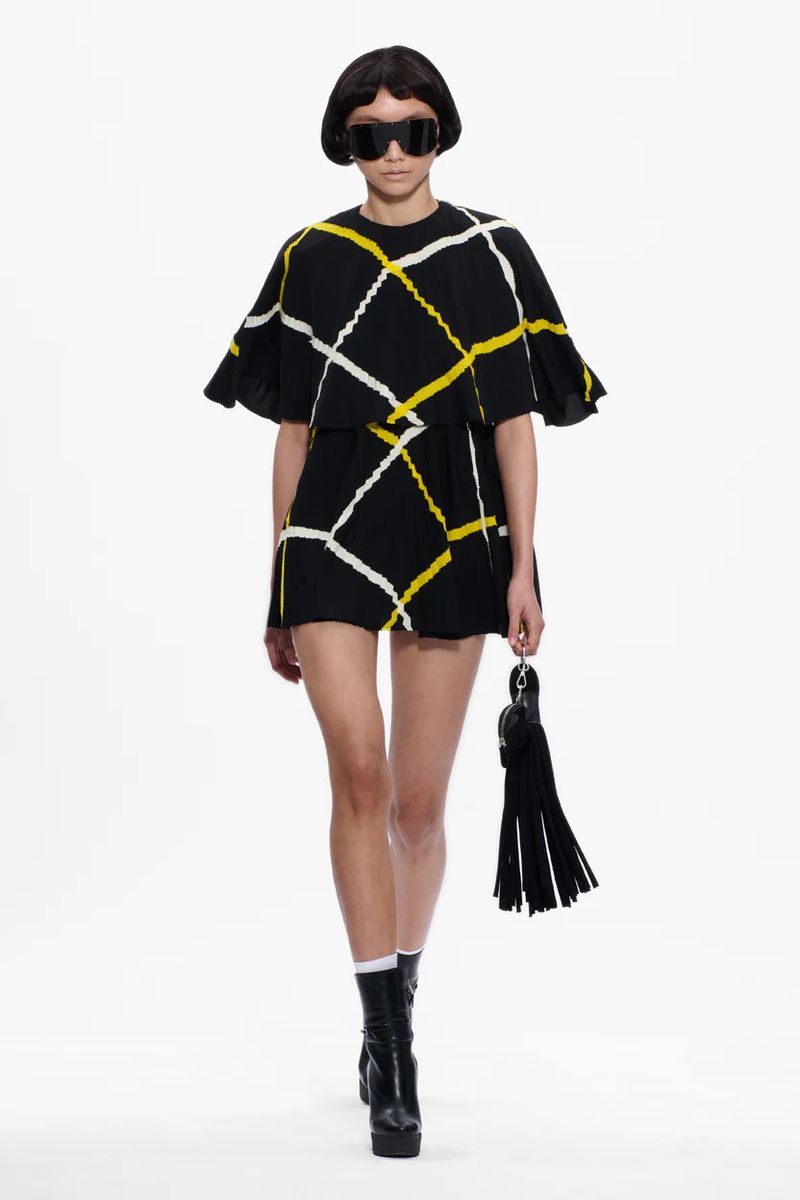 Runway fashion: a model showcases a black dress with white and yellow stripes, paired with platform boots against a white backdrop.