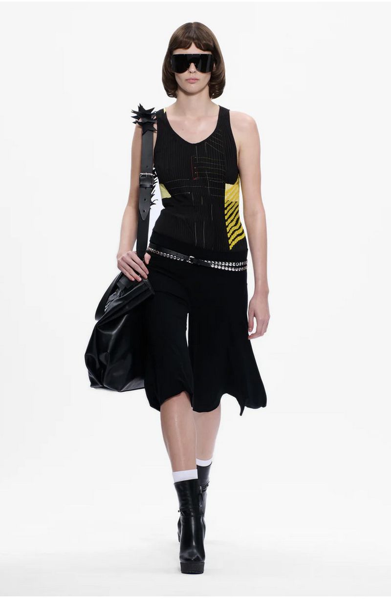 Woman in a black tank top and skirt accessorized with a studded belt and shoulder bag poses in a full shot against a white backdrop.
