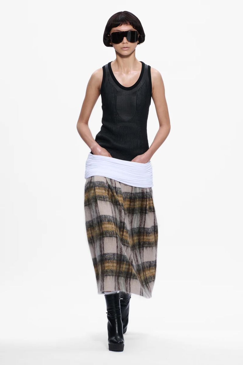 A fashion model walks on a white runway wearing a black tank top, plaid skirt, platform boots, and large sunglasses for a fashion show.