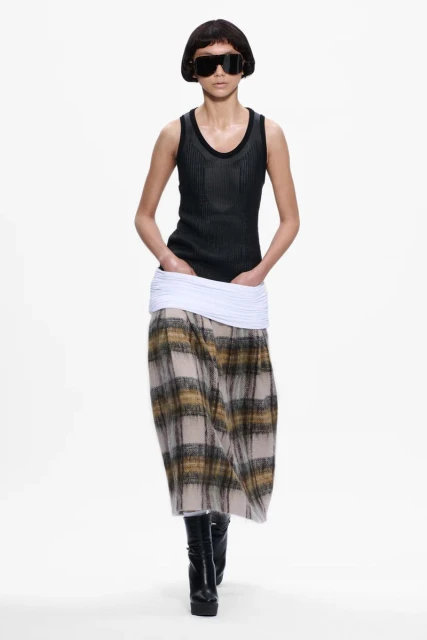 A fashion model walks on a white runway wearing a black tank top, plaid skirt, platform boots, and large sunglasses for a fashion show.