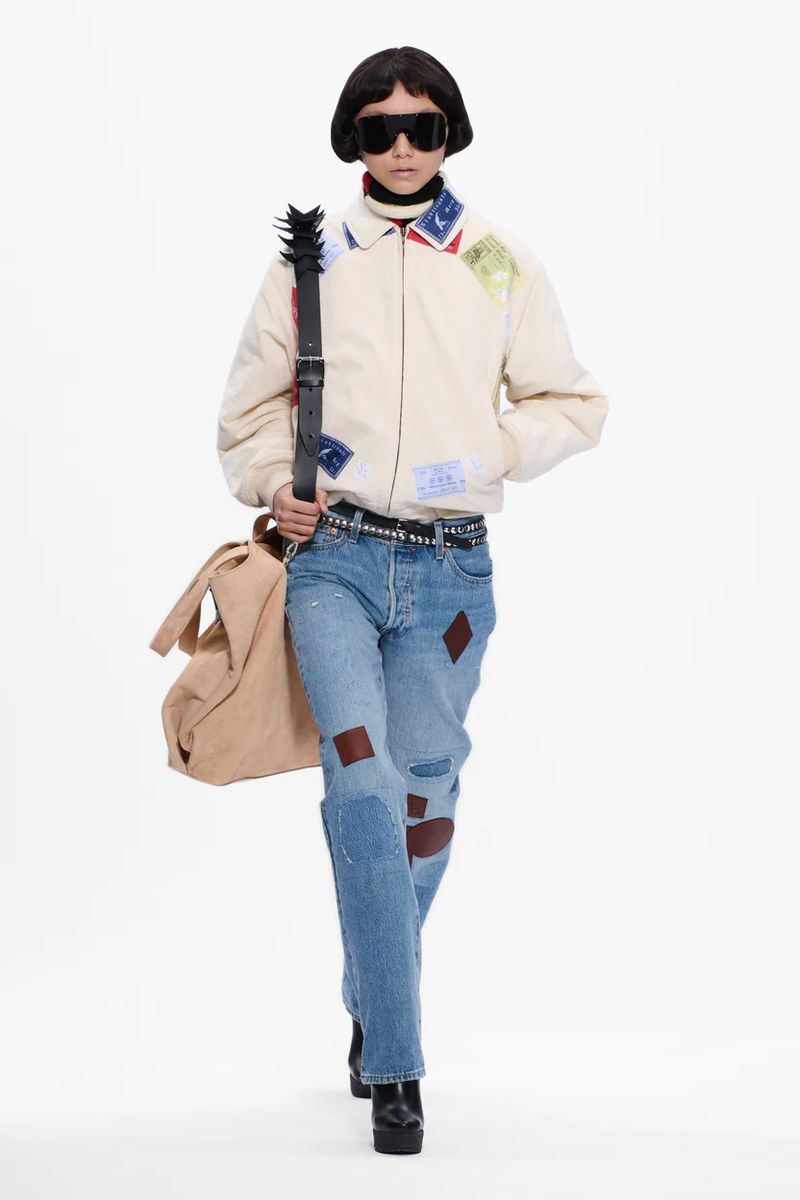 Runway fashion model in a beige patchwork jacket, blue jeans with patches, and black boots on a white backdrop, carrying a beige bag.