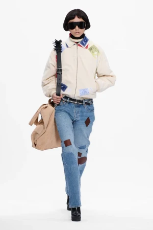 Runway fashion model in a beige patchwork jacket, blue jeans with patches, and black boots on a white backdrop, carrying a beige bag.