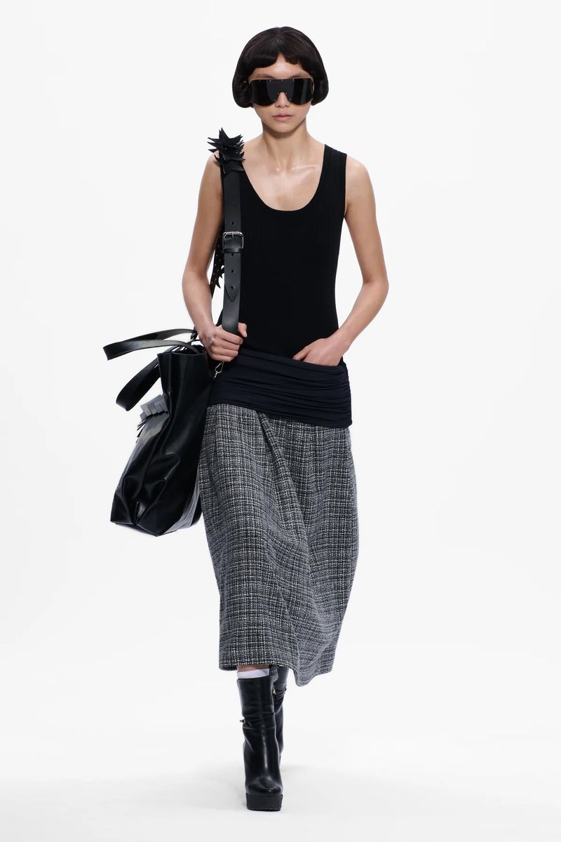 Model wearing dark sunglasses, a black top, and a tweed skirt carrying a large black handbag against a white background
