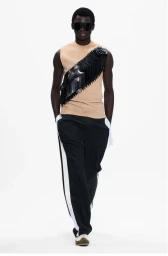 Model wearing dark sunglasses, a beige sleeveless top with black disc embellishment, and black trousers on white seamless background