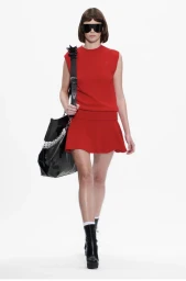 Model in red sleeveless top and mini skirt wearing oversized sunglasses and carrying a black bag against a white seamless background