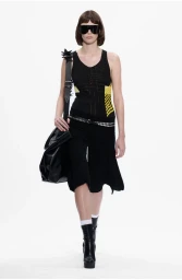 Model wearing dark sunglasses, a black patterned tank top, and black skirt holding a large black handbag against a white background