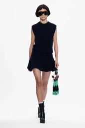 Model in a dark sleeveless top and short skirt walking against a plain white background