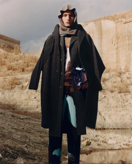 Man in oversized dark coat with hood poses in a desert-like setting in a fashion editorial shot, showcasing unique clothing design.