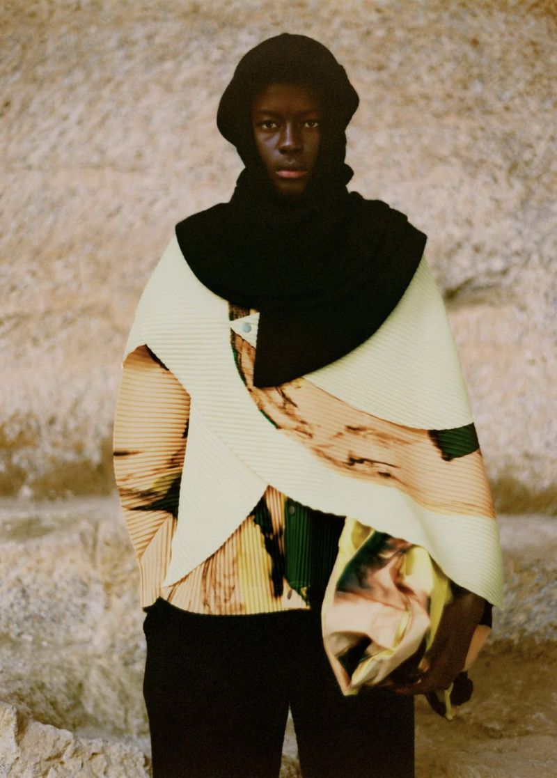 Fashion portrait of an African-descent adult male wearing a black hooded scarf, patterned top, and black trousers against a textured wall.