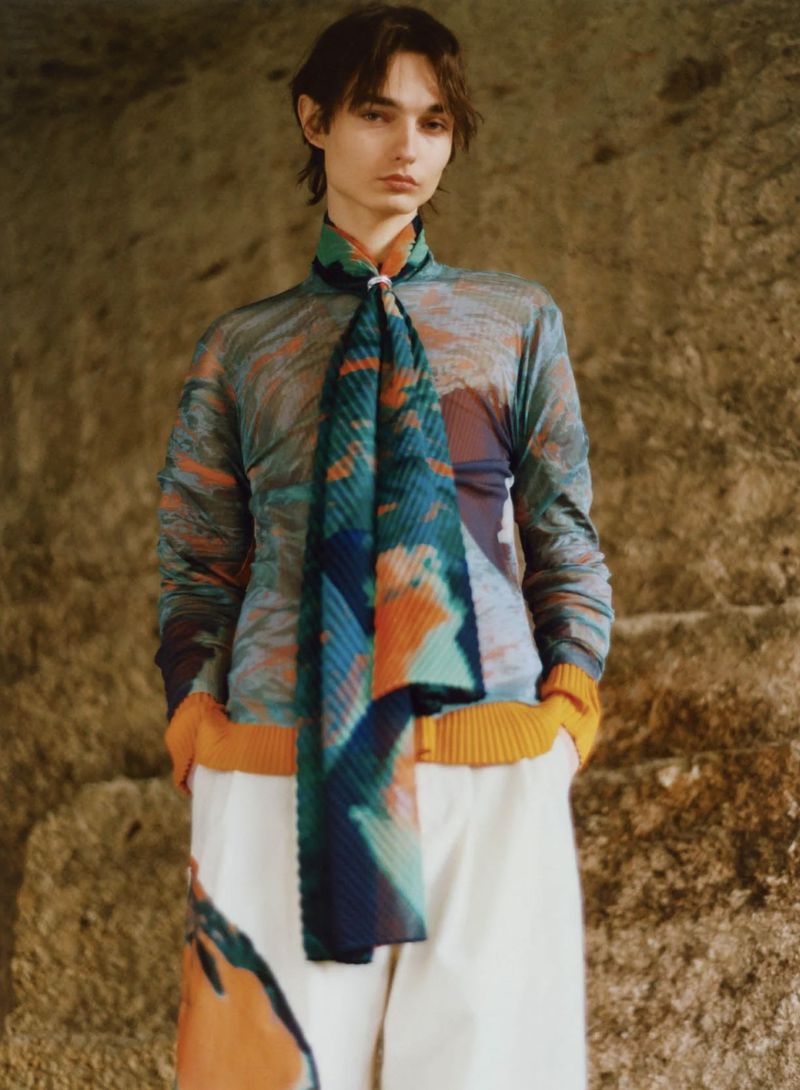 Adult male fashion model in a long-sleeve patterned shirt, wide white pants, and colorful scarf stands indoors against a textured wall.