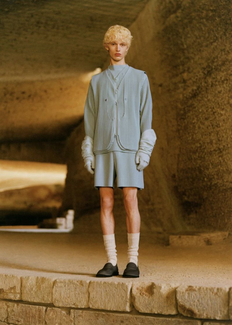A person with blond hair poses on a stone surface in a light blue outfit consisting of a top, shorts, gloves, socks, and shoes.