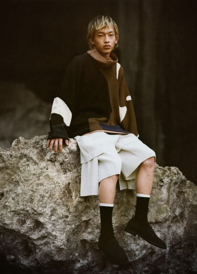 Adult male with blond hair wearing sweater and shorts sitting on rock formation, looking forward in an outdoor environment.