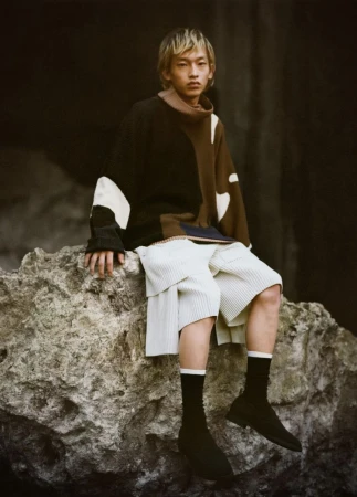 Adult male with blond hair wearing sweater and shorts sitting on rock formation, looking forward in an outdoor environment.