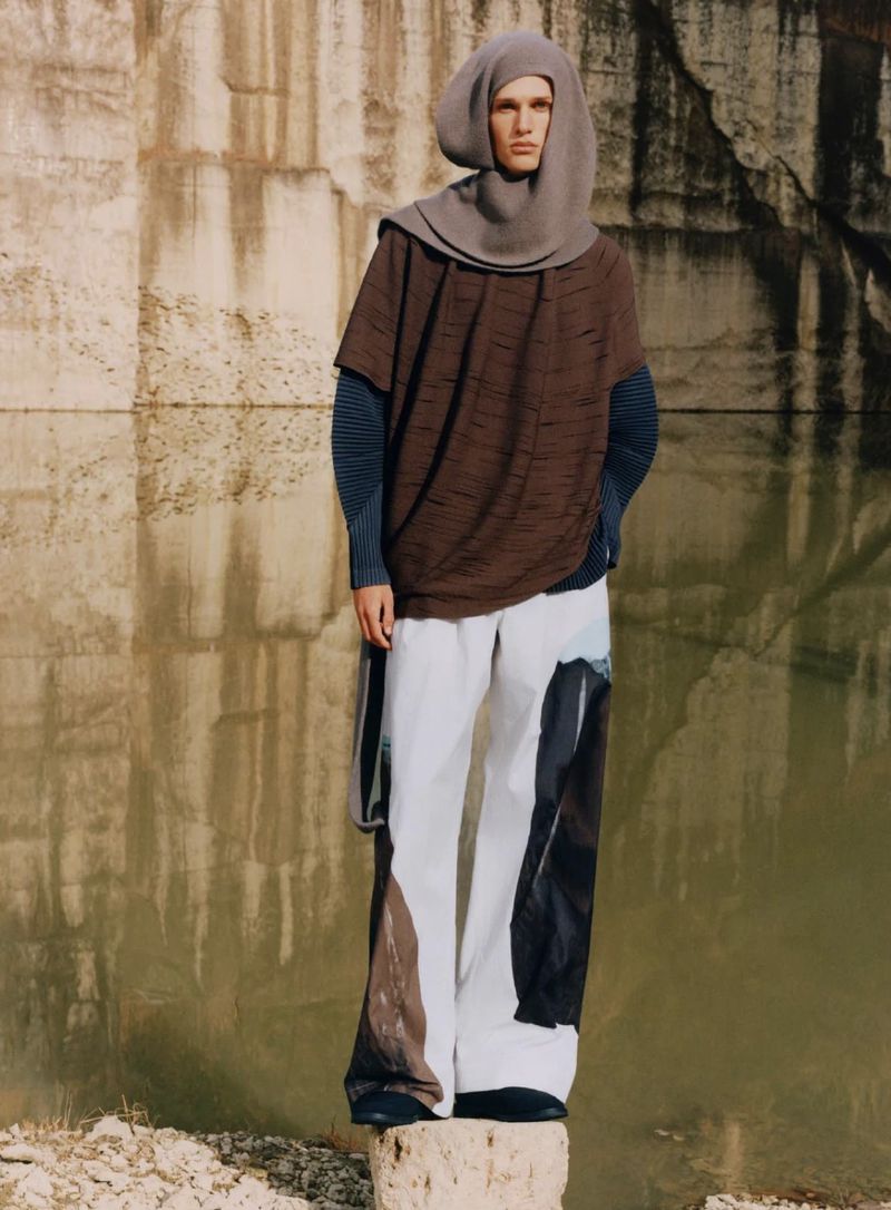 Man stands on rock wearing a hooded poncho, a blue long-sleeve shirt, wide white pants with colored patches, in front of concrete wall.