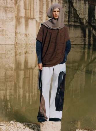 Man stands on rock wearing a hooded poncho, a blue long-sleeve shirt, wide white pants with colored patches, in front of concrete wall.