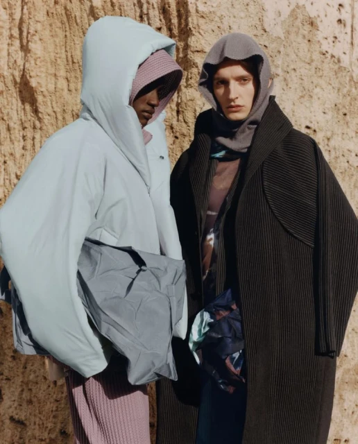 Two models in hooded outerwear pose against a textured brown wall, showcasing fashion with a minimalist and earthy aesthetic in a studio setting.