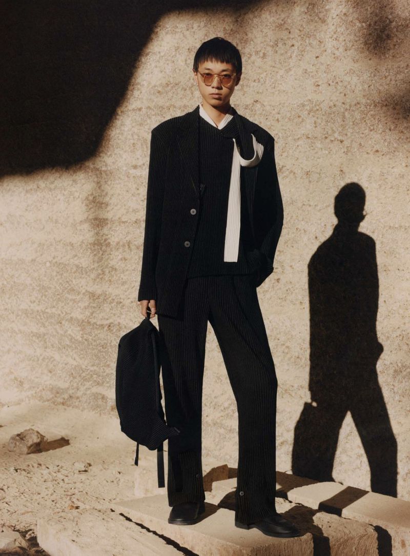 Asian man in a black suit with glasses stands outside on stone blocks, carrying a black backpack, creating a fashion-editorial image.
