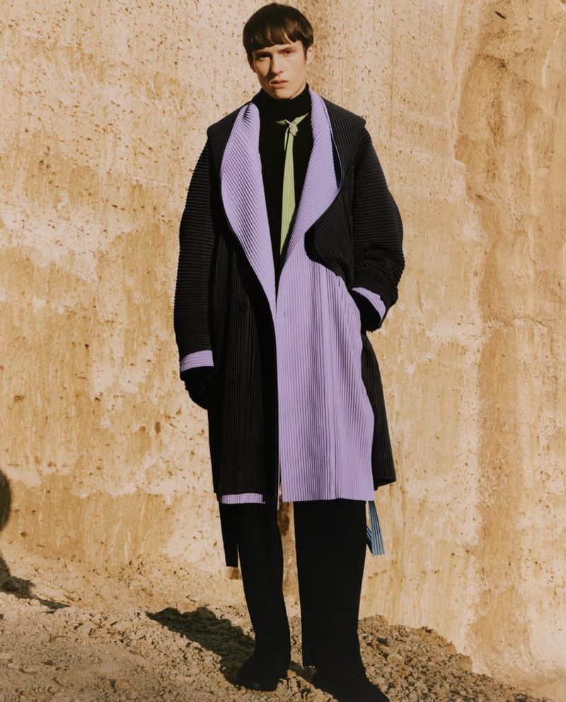 A model wearing a black and lavender overcoat poses in front of a textured wall in a studio setting for a fashion editorial shot.