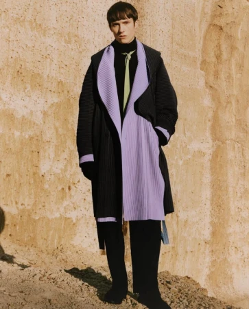 A model wearing a black and lavender overcoat poses in front of a textured wall in a studio setting for a fashion editorial shot.