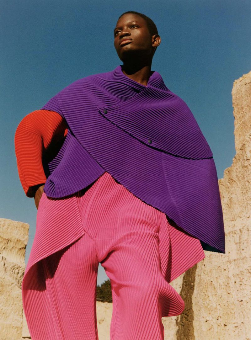 Androgynous person with dark skin poses in avant-garde pink pants and purple cape against tan rock and blue sky in outdoor fashion shot.