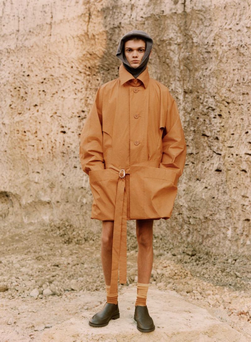 Full-length shot of a young adult male posing outdoors, wearing an orange belted trench coat with a gray balaclava and black shoes.