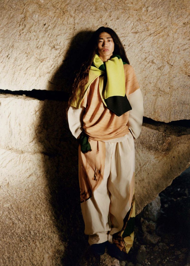 Adult male stands in front of a stone wall wearing cream-colored pants, tan shirt, and green and yellow scarf in a studio setting.