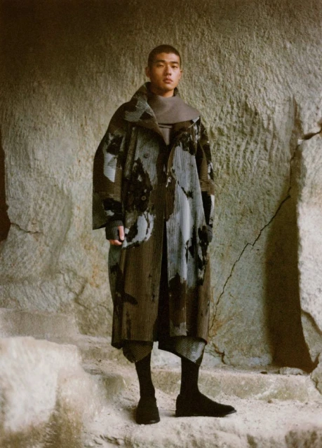 An adult male stands in front of a rock wall wearing a long, patterned coat, dark socks, and shoes in a neutral and earthy color palette.