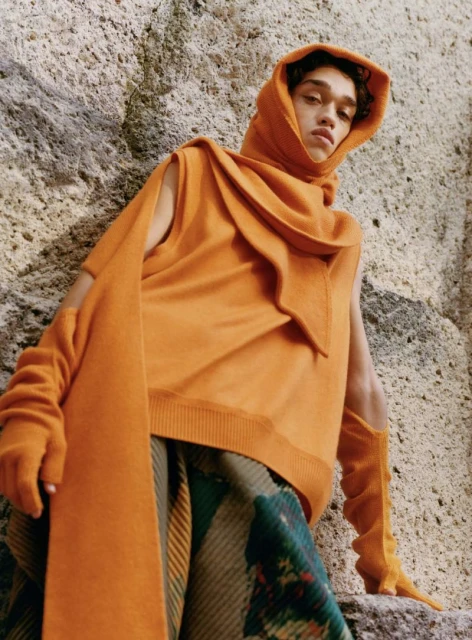 A young man poses outdoors wearing an orange hooded sweater, scarf, and gloves, with patterned trousers and textured stone background.