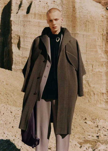Fashion portrait of a young adult male model wearing an oversized striped overcoat and pleated pants standing in a desert-like environment.