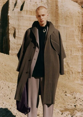 Fashion portrait of a young adult male model wearing an oversized striped overcoat and pleated pants standing in a desert-like environment.