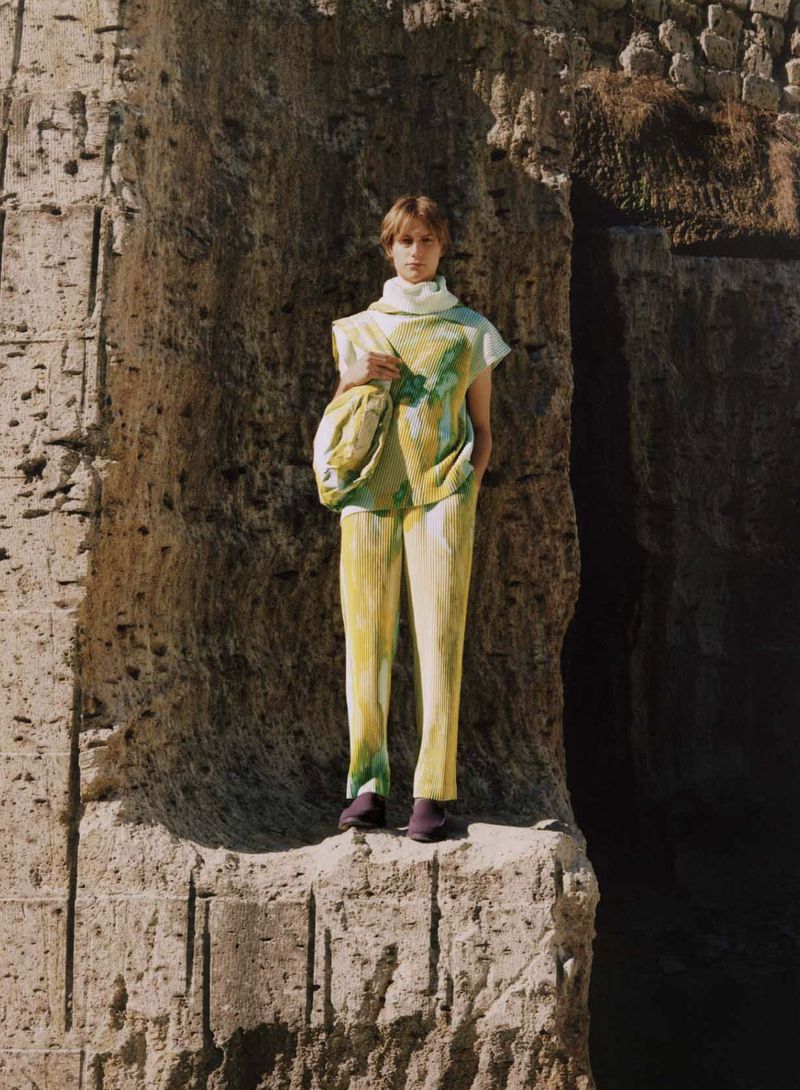 Adult female fashion model in an outdoors urban environment wearing yellow and green matching outfit with shoulder bag standing in front of stone wall.