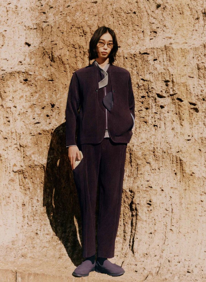 A stylish adult male model in a textured purple suit, posing outdoors against a textured brown wall in soft natural light.