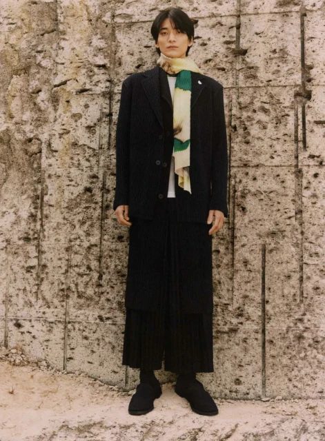 An adult male is standing in front of a textured wall wearing a dark blazer, trousers, a scarf, and shoes in a full shot.