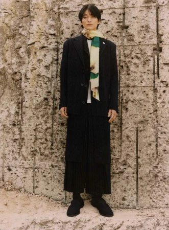 An adult male is standing in front of a textured wall wearing a dark blazer, trousers, a scarf, and shoes in a full shot.