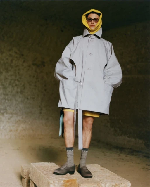 Man in oversized grey coat and yellow scarf stands on stone blocks in an eroded stone environment. Fashion and portrait photography.
