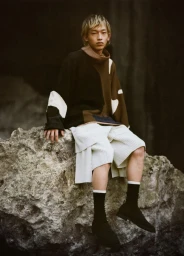Model seated on a rocky outcrop wearing a colorblocked sweater and textured white shorts for Issey Miyake.