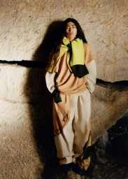 Long-haired man wearing layered knitwear and patterned trousers standing against a textured stone wall