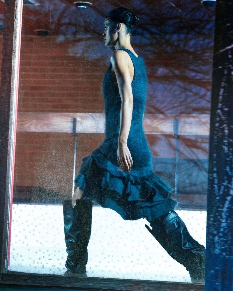 Woman poses in a blue ruffled dress and black boots in a studio with a brick background and city reflection in the foreground.