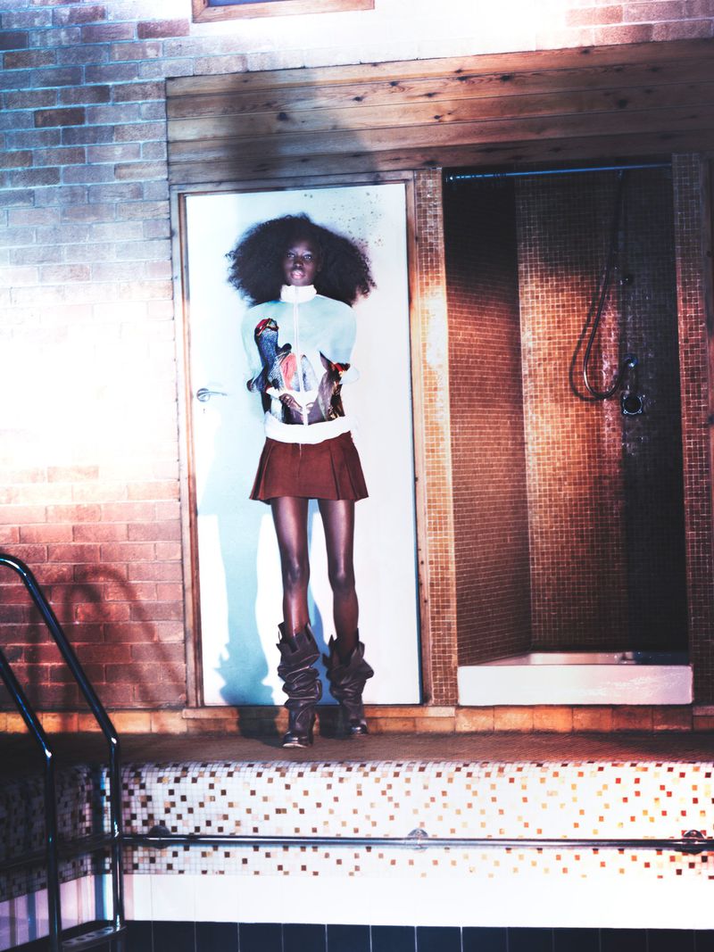 Fashion shot of a slender Black woman with an afro, wearing a retro-style mini skirt, sweater, and high boots standing by an indoor pool.