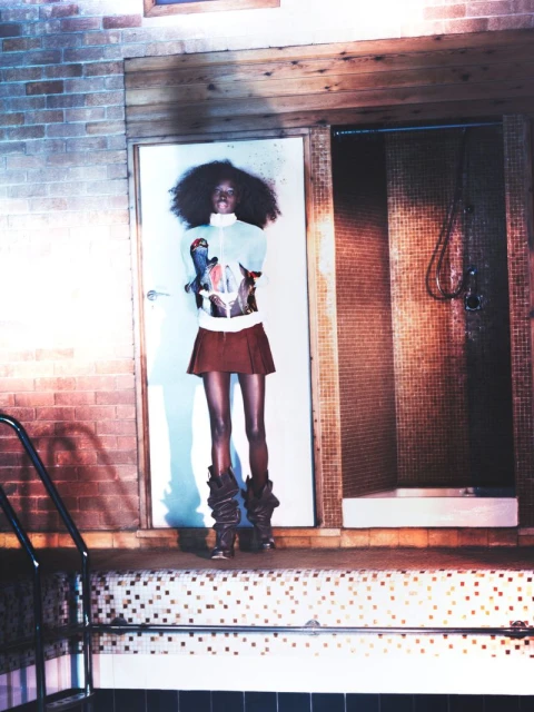 Fashion shot of a slender Black woman with an afro, wearing a retro-style mini skirt, sweater, and high boots standing by an indoor pool.