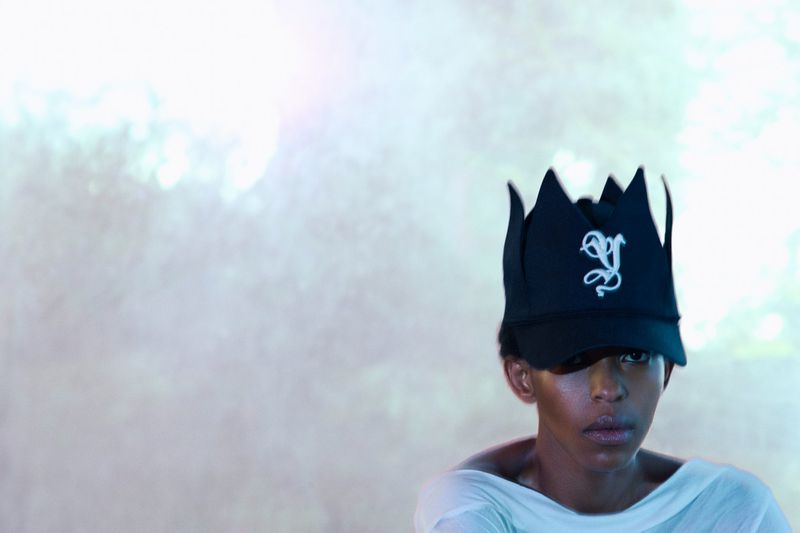Person wears a black crown baseball cap with an embroidered design, posing against a soft, blurred background with gradient lighting.
