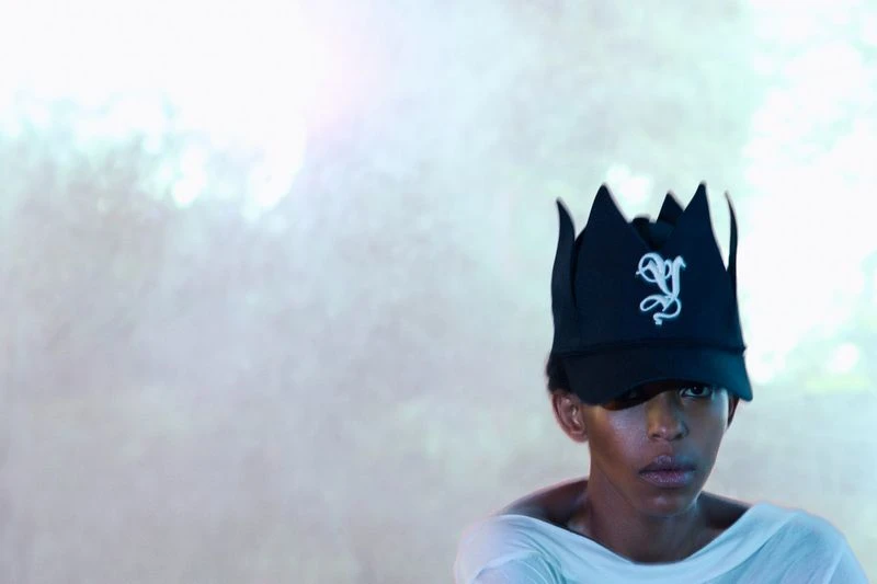 Person wears a black crown baseball cap with an embroidered design, posing against a soft, blurred background with gradient lighting.