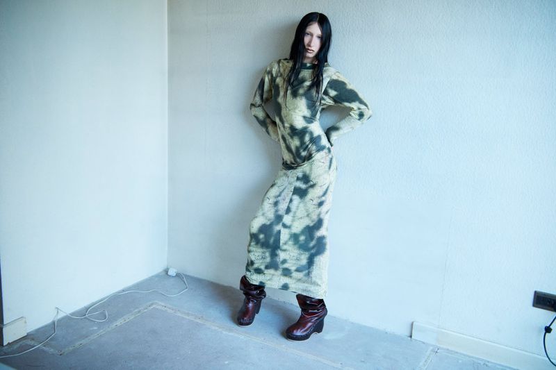 Woman in tie-dye dress and platform boots poses in a corner of an empty room with bare walls and a concrete floor, looking at the camera.